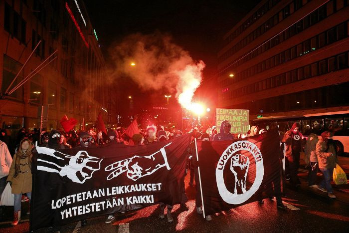 Na transparentu levo piše: "zaustavimo rezove (nap. prev. mere štednje), kolektivizujmo sve". Na transparentu desno piše "klasni rat, sada".